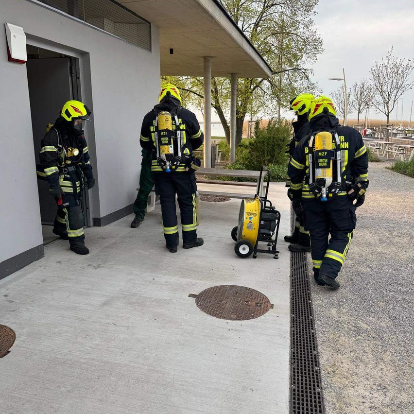 Feuerwehrleute in Schutzkleidung stehen am Eingang eines Gebäudes, wobei ein Mann eine Sauerstoffflasche trägt. Dahinter befindet sich eine gelbe Maschine auf einem Wagen.