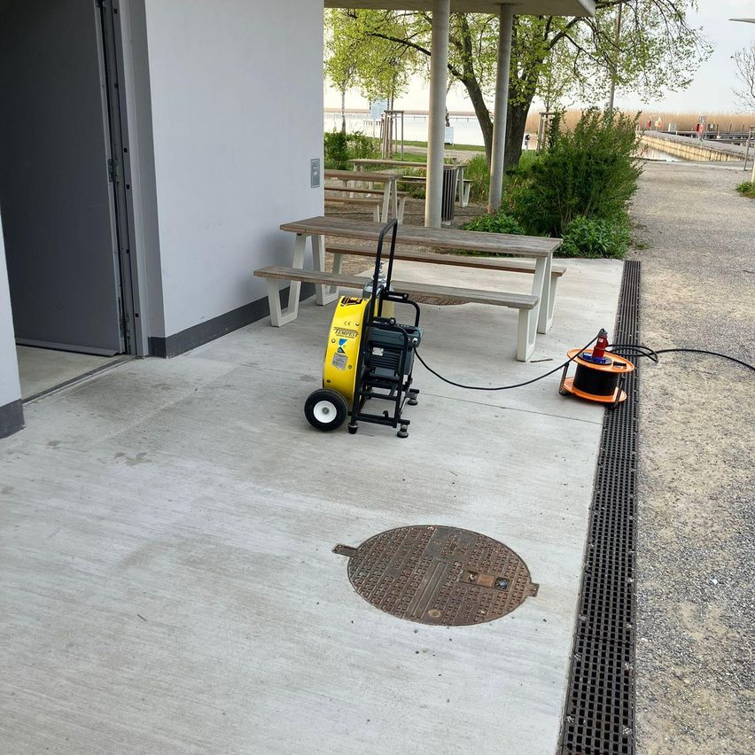 Eine gelbe und schwarze Maschine steht auf einer Betonplattform neben einer Bank und einem Baum. Es gibt einen Schachtdeckel auf dem Boden.