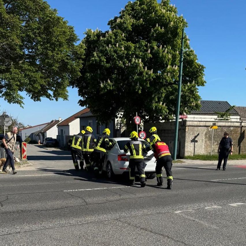 Rettungskräfte helfen jemandem aus einem beschädigten Auto auf einer Straße. Bäume und Gebäude sind im Hintergrund.