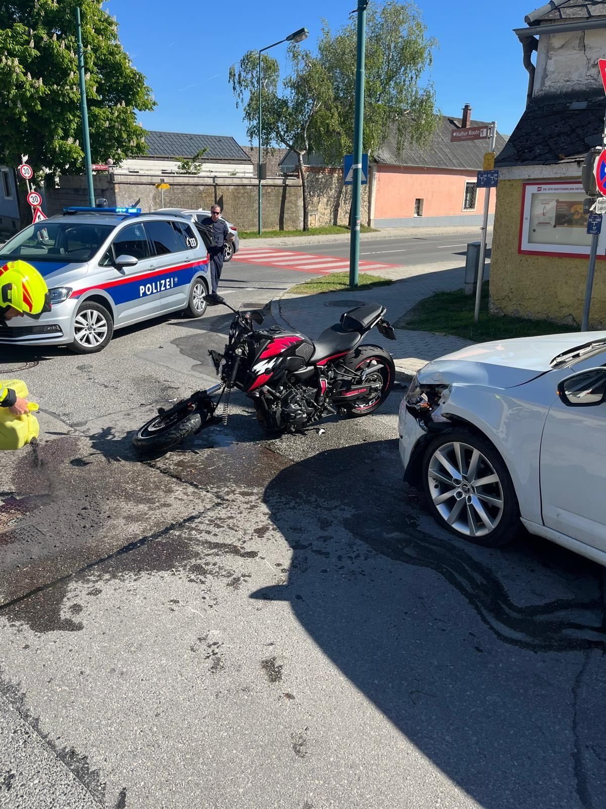 Ein Motorrad und ein Auto sind beschädigt auf einer Straße. Ein Polizeibeamter ist anwesend, und eine Person in einer gelben Sicherheitsweste ist in der Nähe.