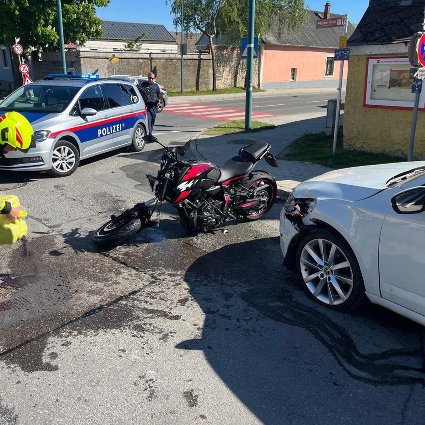 Ein Motorrad und ein Auto sind beschädigt auf einer Straße. Ein Polizeibeamter ist anwesend, und eine Person in einer gelben Sicherheitsweste ist in der Nähe.