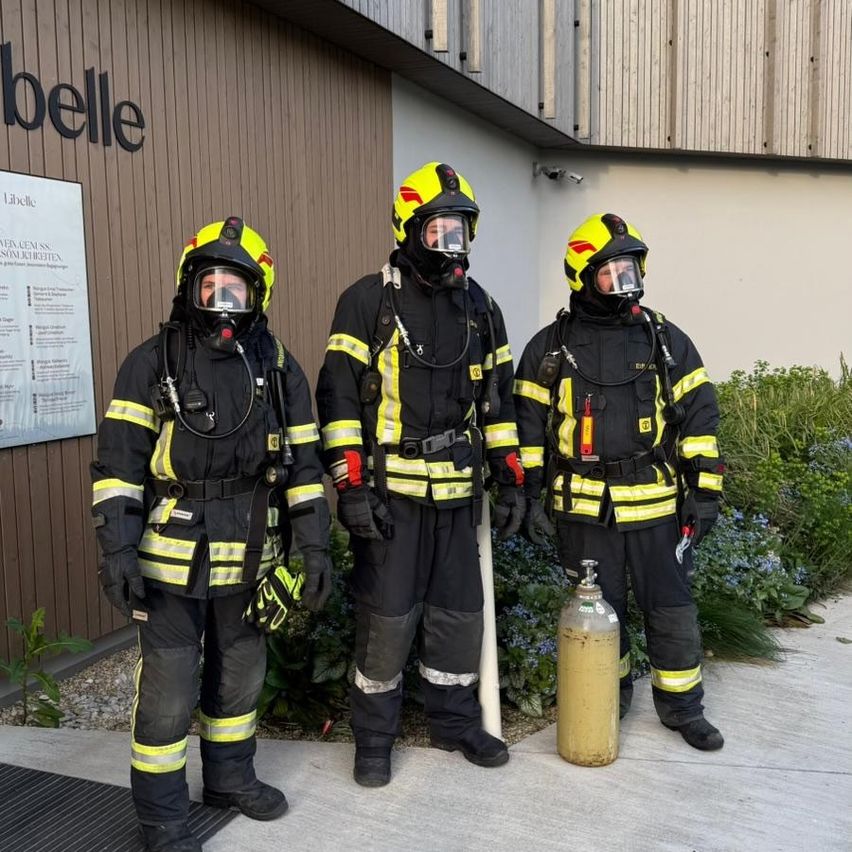 Drei Feuerwehrleute in voller Ausrüstung stehen draußen neben einem Feuerlöscher. Sie stehen vor einem Gebäude mit dem Wort Belle darauf.