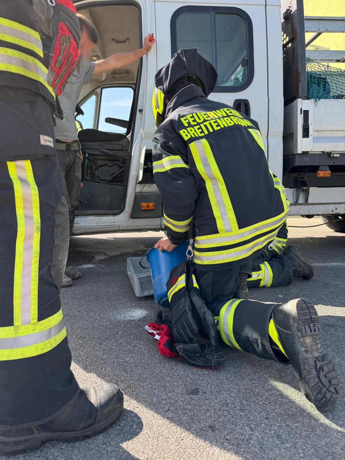 Feuerwehrleute knien neben einem Lkw, einer hält einen blauen Eimer. Sie tragen reflektierende Westen und Schutzausrüstung.