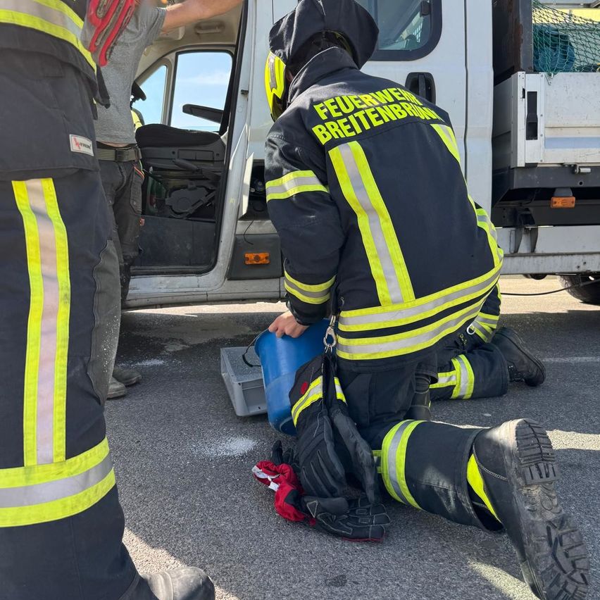 Feuerwehrleute knien neben einem Lkw, einer hält einen blauen Eimer. Sie tragen reflektierende Westen und Schutzausrüstung.