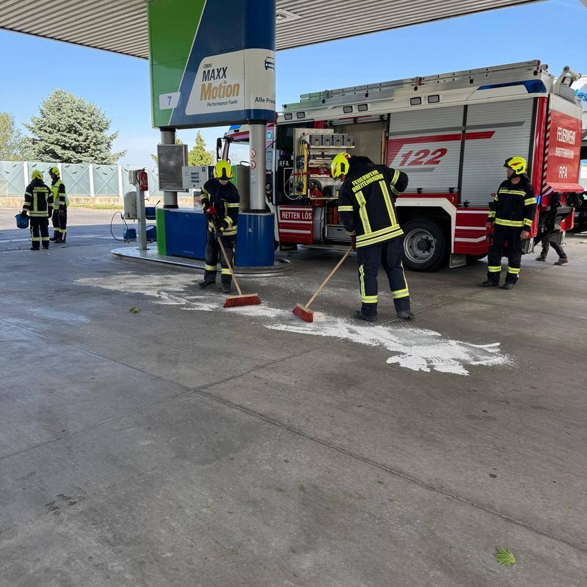 Feuerwehrleute reinigen den Boden an einer Tankstelle mit einem Feuerwehrauto im Hintergrund.