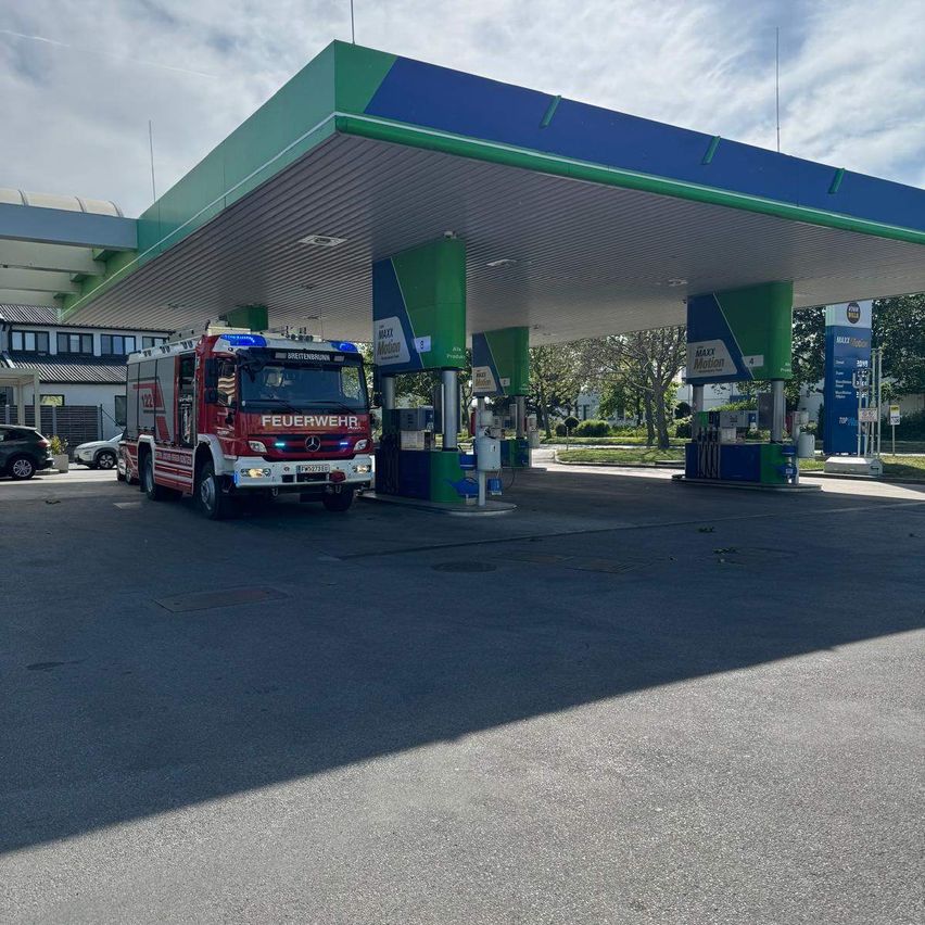 Ein roter Feuerwehrwagen ist an einer Tankstelle geparkt. Er hat den Text 'Feuerwehr' auf der Seite. Rechts sind mehrere Zapfsäulen.