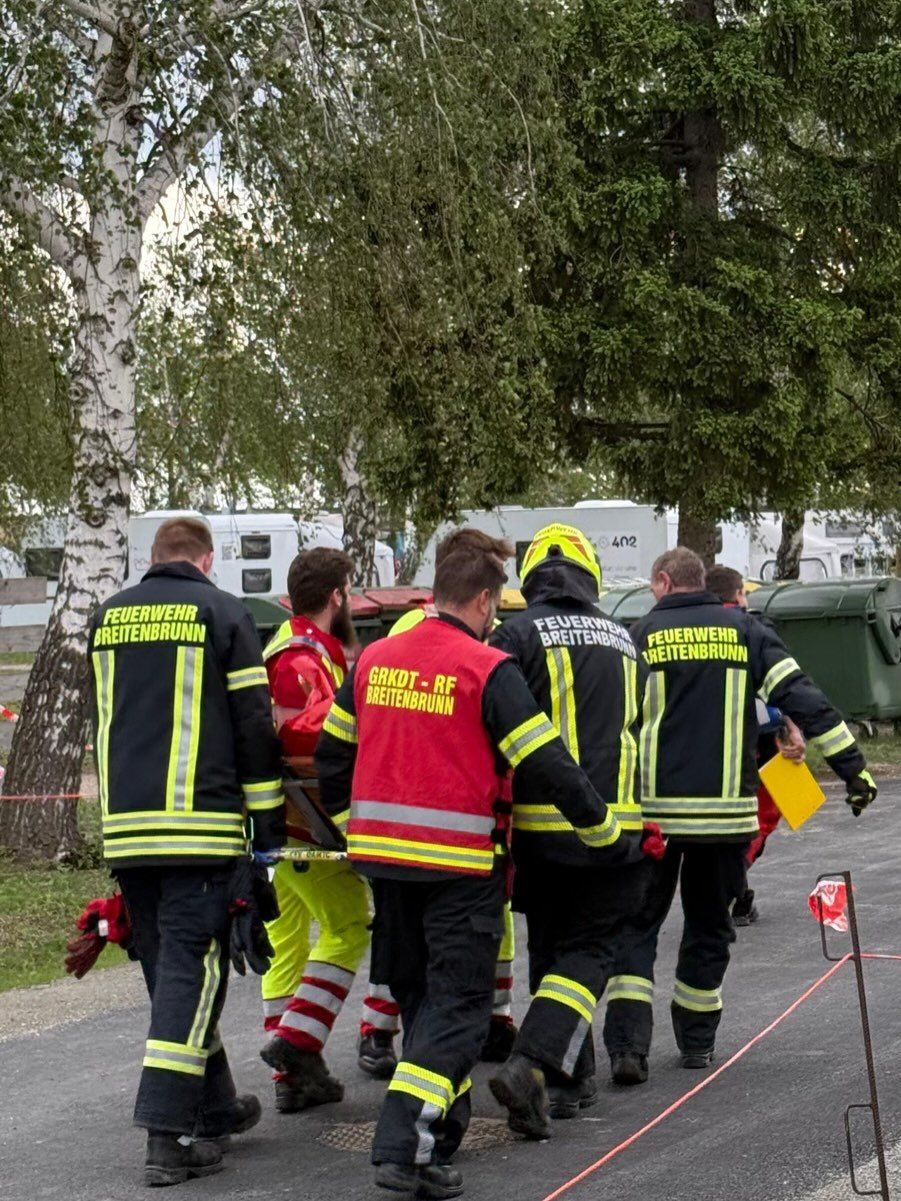 Feuerwehrleute in Uniform tragen eine Trage mit einer Person darauf. Sie sind von Bäumen und einer grünen Mülltonne umgeben.