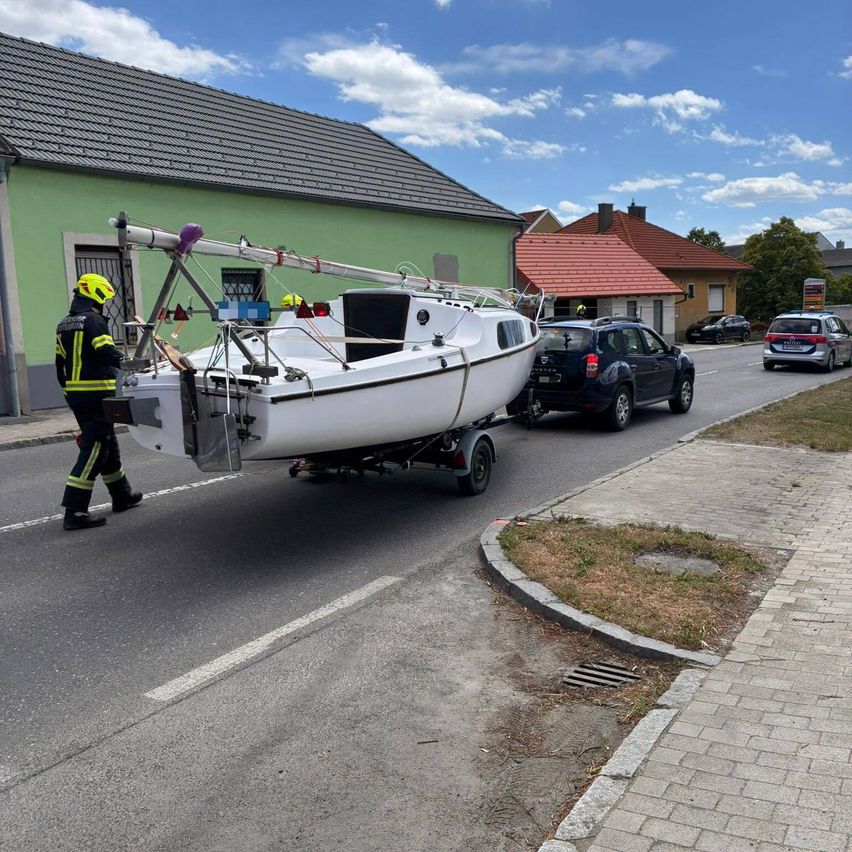 Ein Feuerwehrmann zieht ein Boot mit Anhänger auf einer Straße, mit geparkten Autos und Häusern im Hintergrund.
