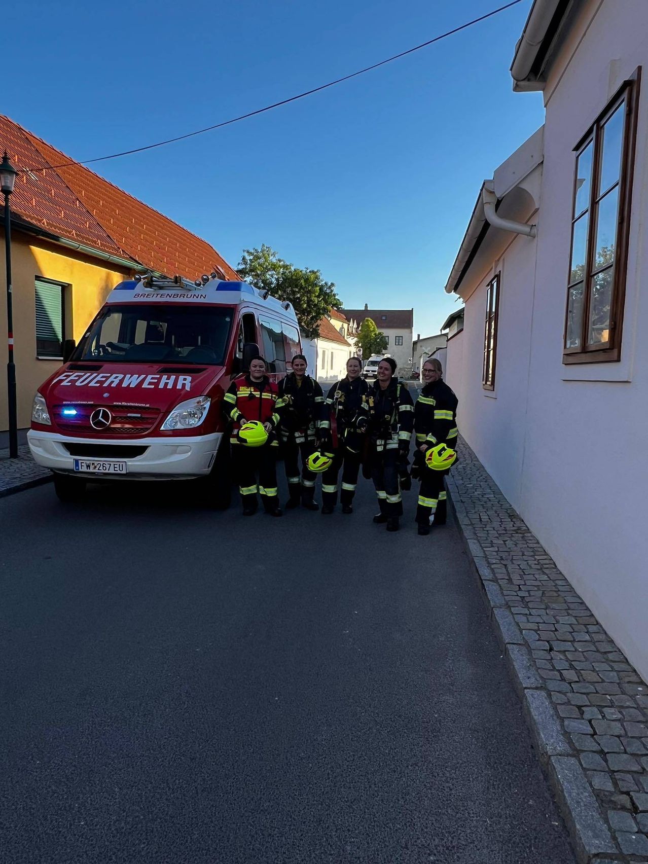 Eine Gruppe Feuerwehrleute steht vor einem Feuerwehrauto, trägt Schutzausrüstung und gelbe Helme, mit einem Gebäude und einem Baum im Hintergrund.