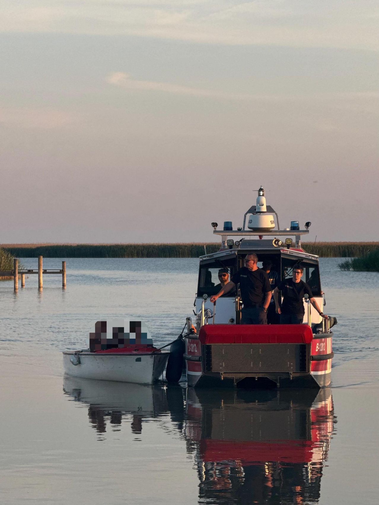 Ein Feuerwehrboot zieht ein kleineres Boot über einen See. Vier Männer sind auf dem Feuerwehrboot, einer von ihnen lenkt.