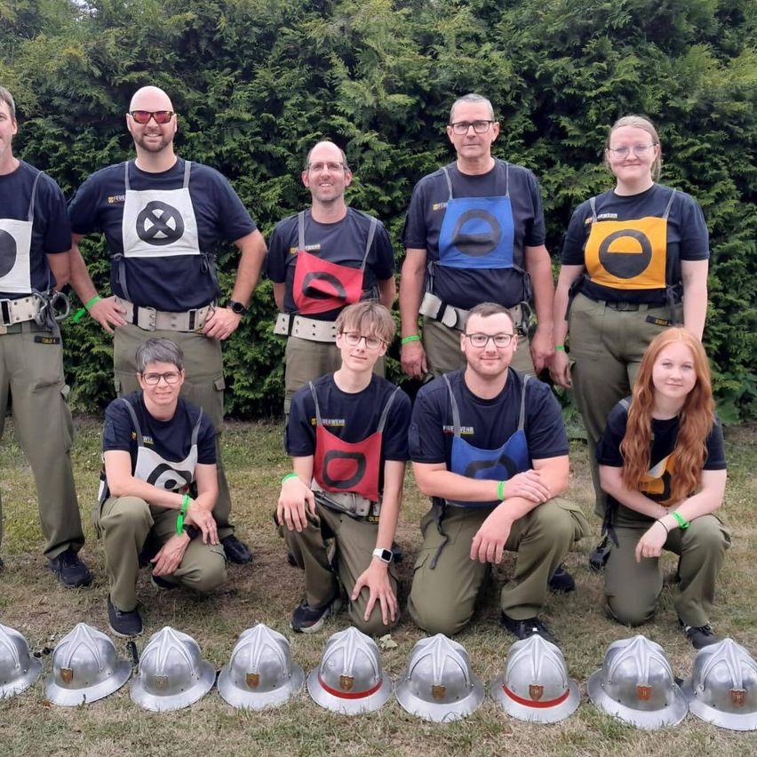 Eine Gruppe von Menschen in grünen Uniformen mit Sicherheitswesten posiert für ein Foto mit Helmen vor ihnen und einem grünen Busch als Hintergrund.
