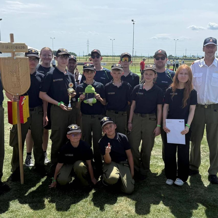 Eine Gruppe junger Personen in Uniformen posiert für ein Foto auf einem Feld mit einem Pokal und einem Holzschild.