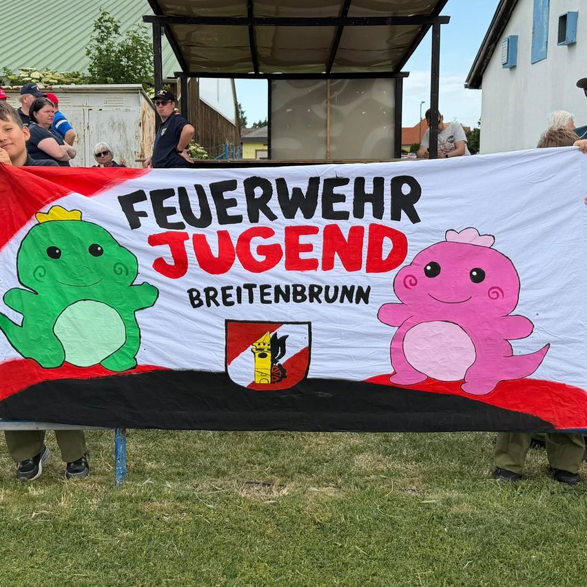 Ein farbenfrohes Banner mit Cartoon-Charakteren und den Worten 'Feuerwehr Jugend Breitenbrunn' wird von zwei Personen bei einer Outdoor-Veranstaltung gehalten. Mehrere Personen beobachten hinter dem Banner.