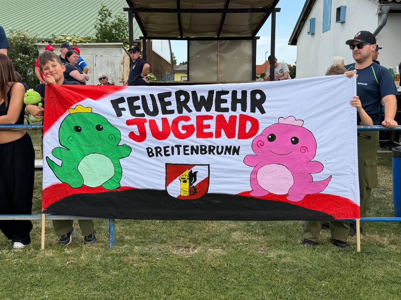 Ein farbenfrohes Banner mit Cartoon-Charakteren und den Worten 'Feuerwehr Jugend Breitenbrunn' wird von zwei Personen bei einer Outdoor-Veranstaltung gehalten. Mehrere Personen beobachten hinter dem Banner.