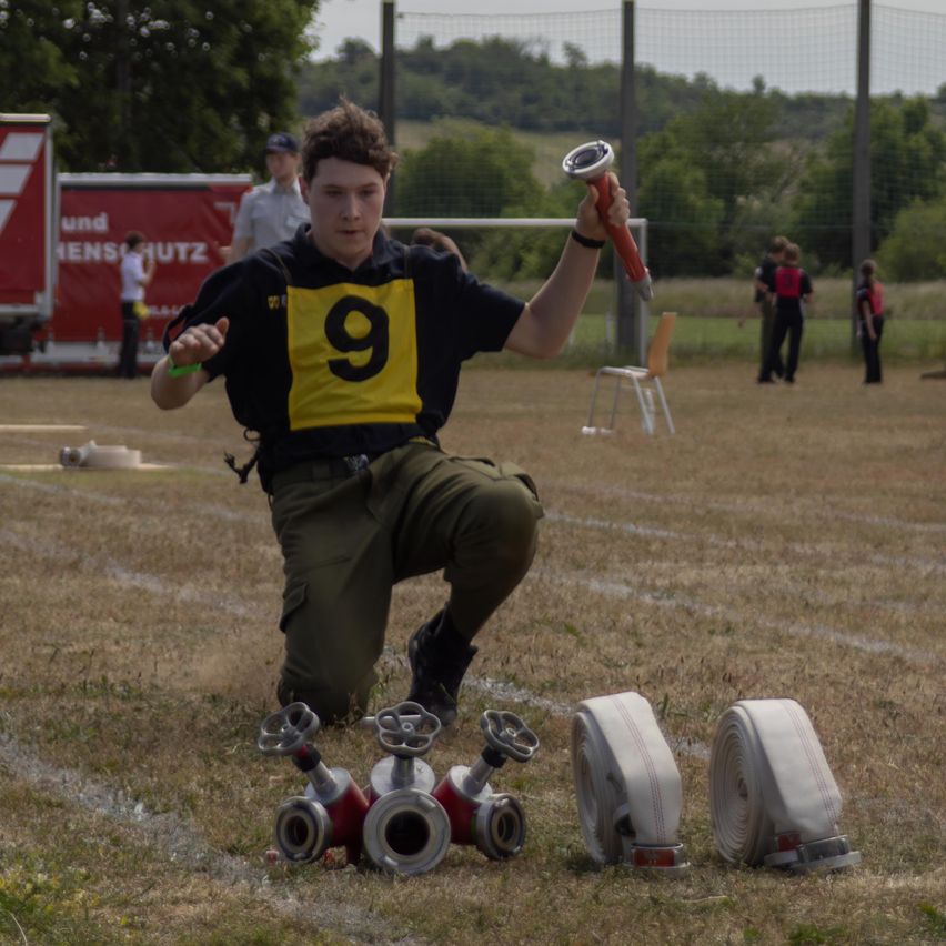 Ein junger Mann kniet auf einem Feld und hält einen Feuerwehrhydrantenschlüssel. Er trägt ein gelbes Shirt mit der Nummer 9. Feuerwehrschläuche und Feuerlöscher liegen um ihn herum. Im Hintergrund gehen Leute in der Nähe eines roten Lastwagens.