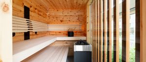 Eine Holzsauna mit Holzbänken, einem schwarzen Eimer, einem Holzkohlegrill und Glasfenstern. Die Holzwände und die Decke sind sichtbar.