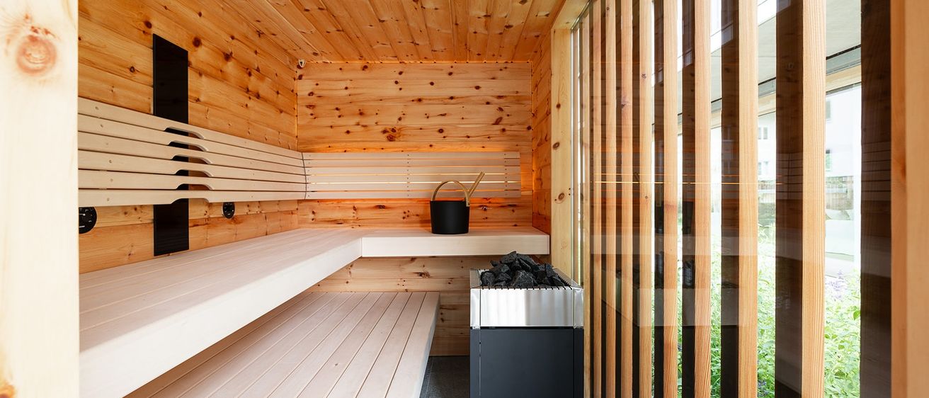 Eine Holzsauna mit Holzbänken, einem schwarzen Eimer, einem Holzkohlegrill und Glasfenstern. Die Holzwände und die Decke sind sichtbar.