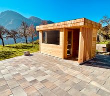 Eine Holzsaunahütte steht auf einer gefliesten Terrasse mit Blick auf Berge und Bäume im Hintergrund.