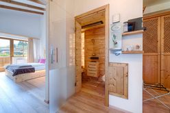 Eine offene Sauna mit hölzerne Innenausstattung, einer Glastür und einer hölzernen Schublade. Daneben ein Schlafzimmer mit einem Bett und einem rosa Kissen.