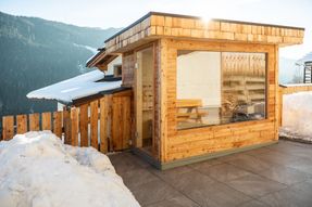 Ein Holzhaus mit Glaswand, schneebedecktem Dach und einem Holzzaun in einem Berggebiet.