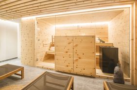 Eine Innensauna mit Holzwänden und einer Glastür. Im Inneren gibt es eine Bank, Handtücher und ein kleines Regal mit einer Pflanze. Daneben steht ein Holzschrank. Davor befindet sich ein Holztisch mit Glasplatte.