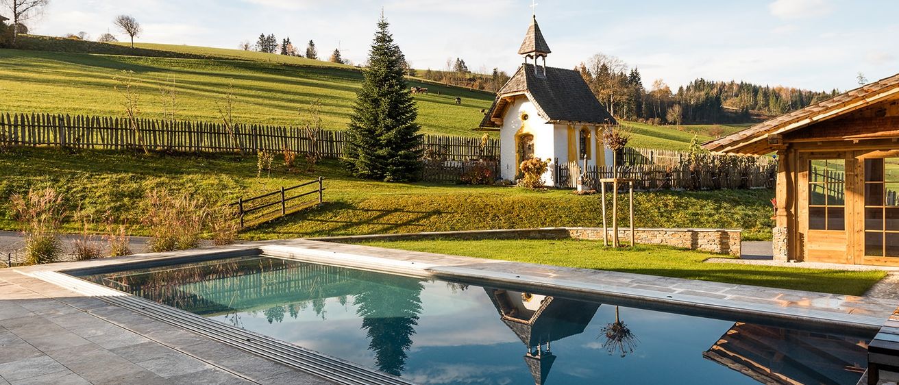 Eine ruhige Kapelle mit einem Turm spiegelt sich in einem klaren Pool, umgeben von einem grasigen Feld und einem Holzzaun unter einem teilweise bewölkten Himmel.