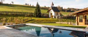 Eine ruhige Kapelle mit einem Turm spiegelt sich in einem klaren Pool, umgeben von einem grasigen Feld und einem Holzzaun unter einem teilweise bewölkten Himmel.