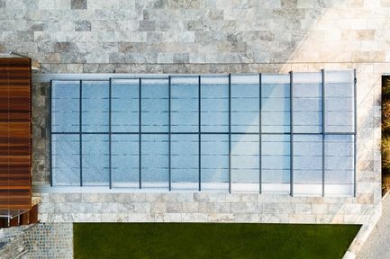 Eine Draufsicht auf einen Swimmingpool mit einem transparenten Glasdach, umgeben von Marmorfliesen und einer grünen Rasenfläche.