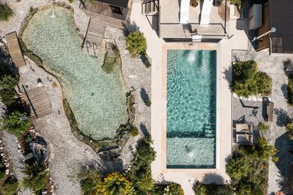 Eine Draufsicht auf ein Grundstück mit zwei Pools, einem größeren und einem kleineren. Der größere Pool hat eine Sprungbrett, und beide sind von Kies und Pflanzen umgeben.