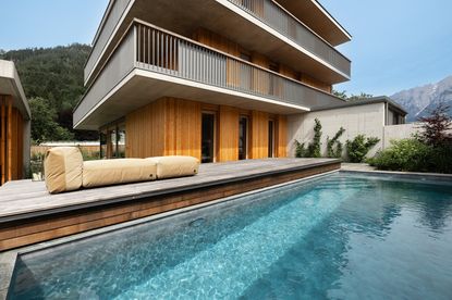 Ein modernes, mehrstöckiges Haus mit holzverkleideten Außenwänden und Balkonen. Das Haus verfügt über einen Pool davor und zwei beige Kissen auf der Terrasse.