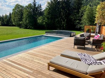 Ein moderner Außenpool mit Glasdach, umgeben von einer Holzterrasse. Liegestühle und ein Tisch sind für Entspannung aufgestellt. Grüner Rasen und Bäume bilden eine ruhige Kulisse.