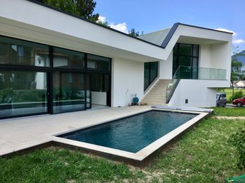 Ein modernes weißes Haus mit Glaswänden und einem Pool davor. Der Pool ist von Gras umgeben.