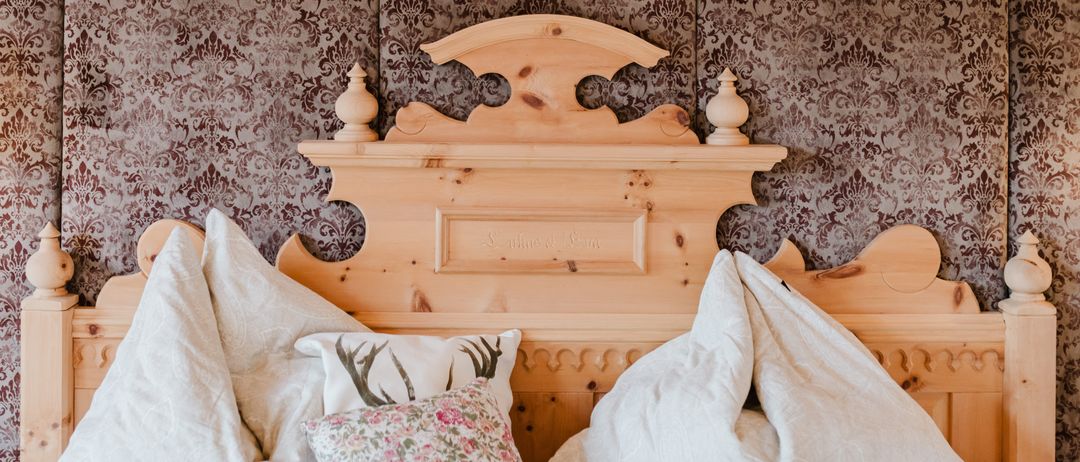 Ein Schlafzimmer mit einem Holzbett und kunstvollem Kopfteil. Das Bett hat weiße Kissen und Kissen mit Blumen und Hirschköpfen. Die Tapete ist braun mit einem Damastmuster.