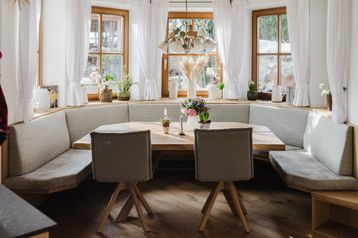 Ein gemütlicher Essbereich mit einem Holztisch, zwei Stühlen und einer Eckbank. Auf dem Tisch stehen Topfpflanzen und Blumen. Der Raum hat Holzböden und weiße Vorhänge.