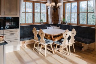 Ein gemütlicher Essbereich mit Holzstühlen und einem Tisch, in einem Raum mit großen Fenstern, Holzwänden und einer Kronleuchter. Der Raum ist von Sonnenlicht durchflutet.