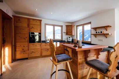 Eine moderne Küche mit Holzschränken, Arbeitsplatten und Bodenbelag. Zwei Hocker stehen neben einer hölzernen Bartheke. Eine Kaffeemaschine und andere Küchenutensilien sind auf der Theke.