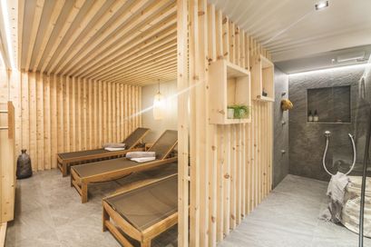 Ein Innenansicht einer Holzsauna mit drei Liegestühlen, einer hängenden Lampe und einem Regal mit einer Pflanze. Der Raum hat Holzwände und -böden.
