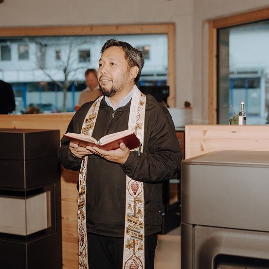 Ein Mann mit einer Priesterkrawatte steht in einem Raum mit einer Holzwand und einem Glasfenster und hält ein Buch. Ein anderer Mann ist im Hintergrund.