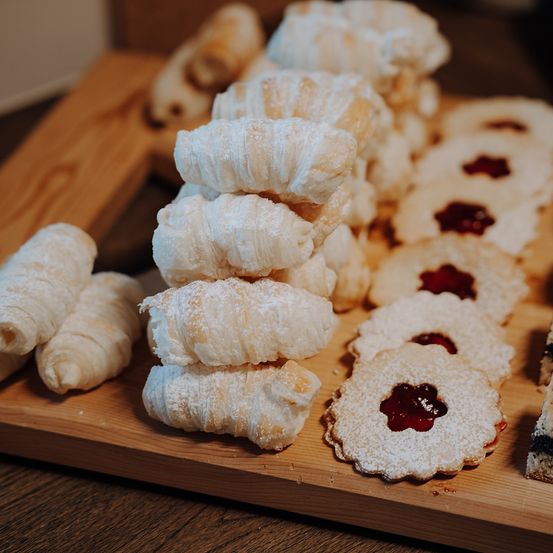 Ein Holzbrett hält einen Stapel Croissants und mehrere Kekse mit Marmeladenfüllungen obenauf.