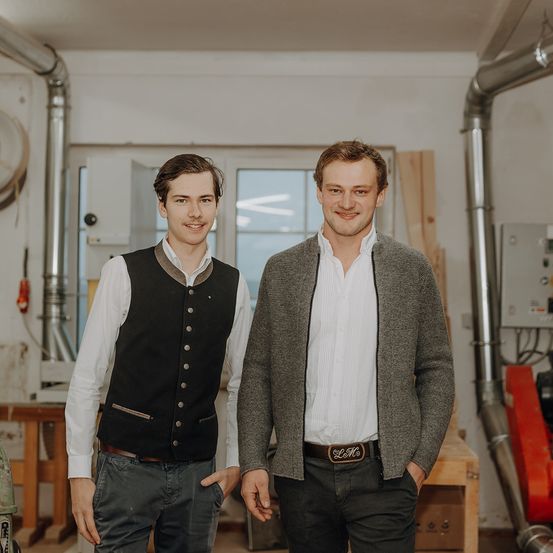 Zwei Männer stehen in einer Werkstatt, einer trägt eine Weste und der andere eine graue Strickjacke. Sie lächeln und scheinen für ein Foto zu posieren. Hinter ihnen befinden sich Holzkonstruktionen, Maschinen und ein Fenster.