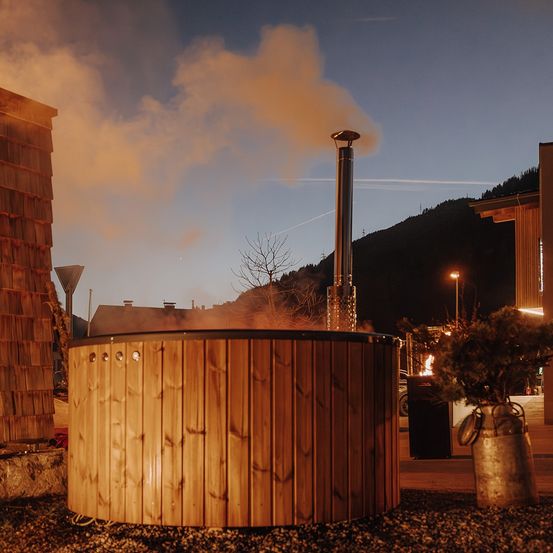 Ein hölzerner Whirlpool dampft bei Dämmerung mit einem Schornstein. In der Nähe ist ein Baum mit einem Feuer beleuchtet. In der Ferne befinden sich Gebäude und Berge.