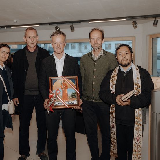 Fünf Personen stehen in einem Raum, ein Mann hält ein gerahmtes religiöses Bild. Sie sind in Winterkleidung gekleidet, ein Mann trägt ein religiöses Gewand. Der Raum hat weiße Wände und Fenster im Hintergrund.