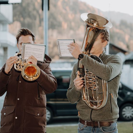 Zwei Männer stehen draußen, einer spielt Trompete und der andere Tuba, beide halten Notenblätter. Dahinter befindet sich ein Gebäude und ein Berg.
