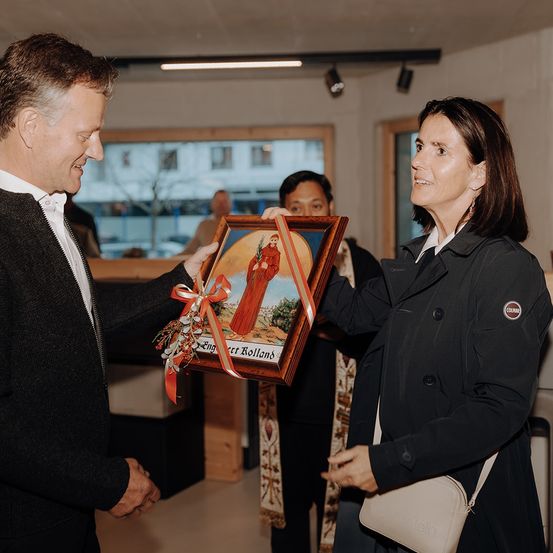Ein Mann und eine Frau stehen sich gegenüber und halten ein gerahmtes Kunstwerk. Das Kunstwerk zeigt einen Heiligen mit einem Band darum. Sie lächeln und scheinen sich in einem zeremoniellen Umfeld zu befinden.