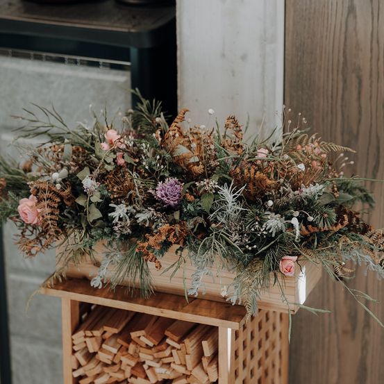 Eine große Blumenarrangement mit getrockneten Blumen, einschließlich rosa, braunen und lila Farbtönen, steht auf einem Holzständer mit einem Holzstapel im Hintergrund.