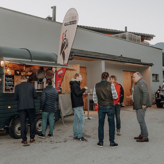Eine Gruppe von Menschen steht vor einem Food Truck, wahrscheinlich um Essen zu bestellen. Dahinter befindet sich ein Gebäude mit einer Fahne.