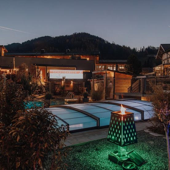 Ein Hotel mit einem Pool und einem Schild mit der Aufschrift 'Schaugarten' wird bei Nacht gesehen. Der Pool ist von einem Glasdach bedeckt. Eine grüne Lampe steht vor dem Pool.