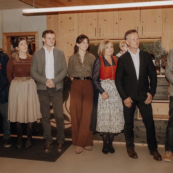 Eine Gruppe von Menschen steht vor einer Holzwand. Sie lächeln und scheinen für ein Foto zu posieren. Dahinter steht ein Holzschrank mit einer Pflanze darauf.
