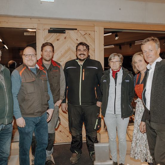 Eine Gruppe von sechs Personen, einschließlich Männer und Frauen, steht vor einer Holzwand, lächelt und posiert für ein Foto. Sie sind warm gekleidet in Jacken und einige tragen Brillen.