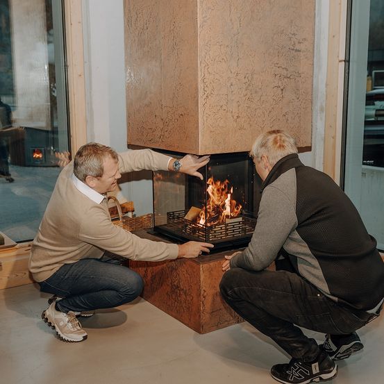 Zwei Männer knien vor einem Kamin, einer zeigt auf die Flammen. Sie sind lässig gekleidet, einer trägt eine Uhr. Glastüren reflektieren das Innere.